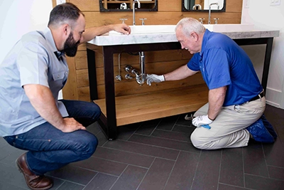 Two men inspecting plumbing under a bathroom sink.