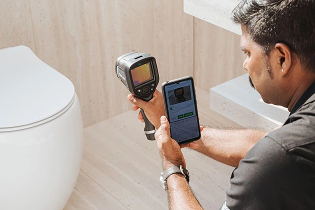 Man using thermal imaging camera near a toilet while checking a smartphone.